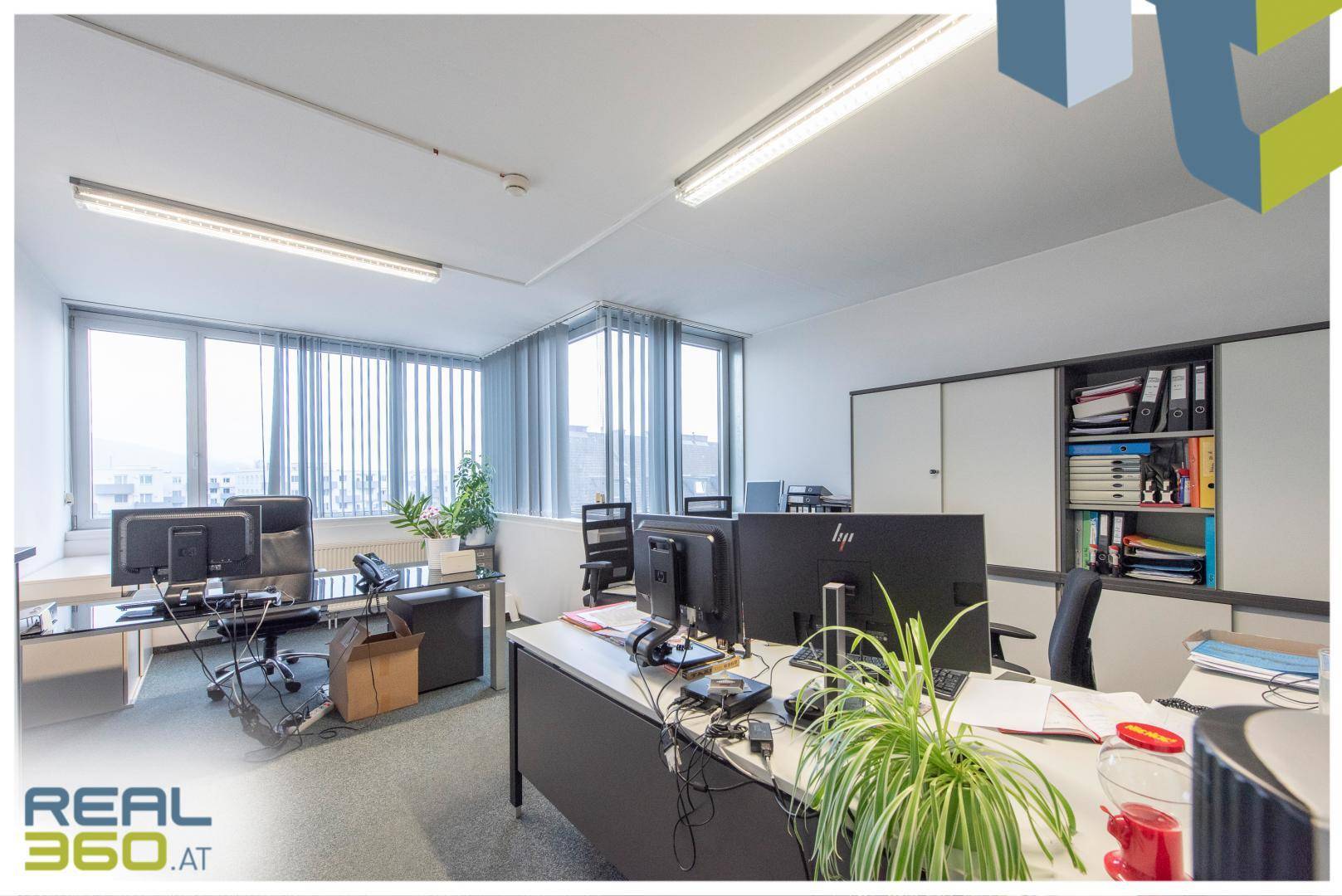 Helles Büro mit mehreren Arbeitsplätzen, großen Fenstern und vertikalen Jalousien für Privatsphäre und Tageslicht.