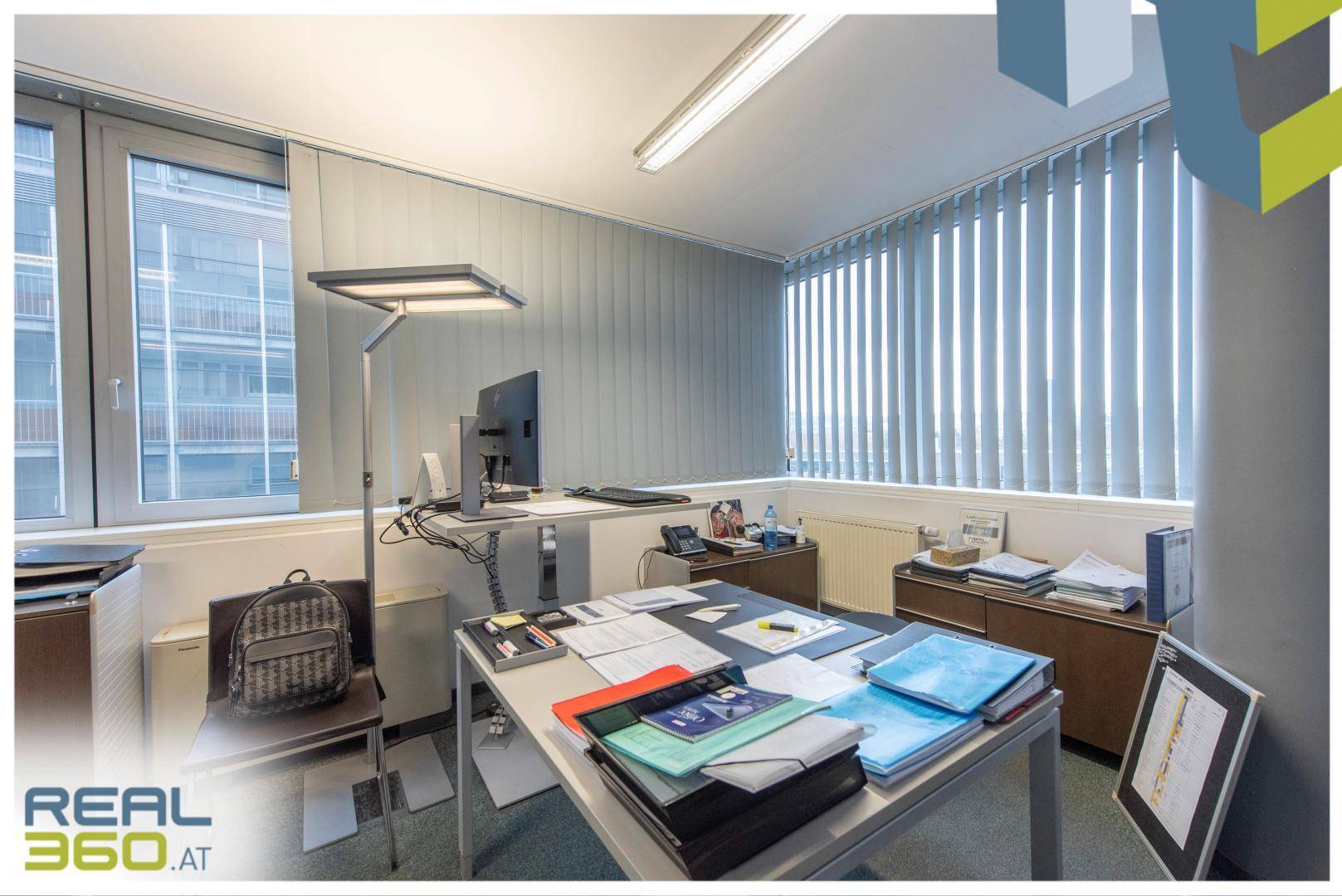 Helles Büro mit Schreibtisch, Dokumenten und großen Fenstern, die viel Tageslicht hereinlassen und Aussicht bieten.