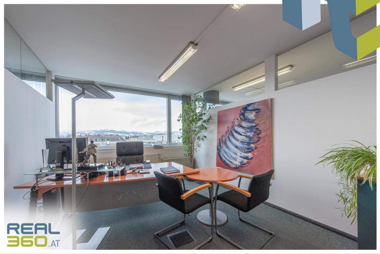 Büro mit modernem Schreibtisch, Kunstwerk an der Wand und großen Fenstern mit Blick auf die Umgebung.