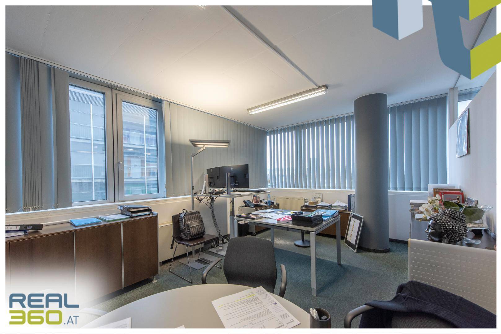 Büro mit mehreren Arbeitsplätzen, einem runden Säulenpfeiler und vertikalen Jalousien.