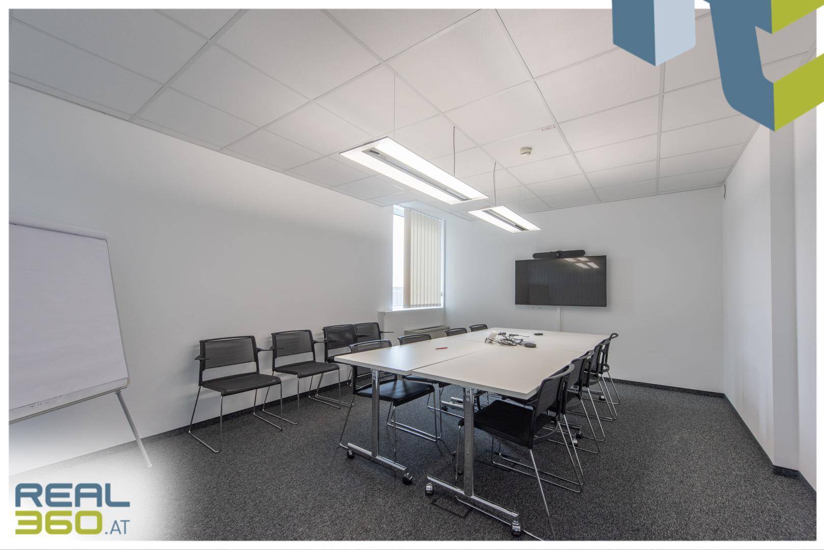 Schlichter Konferenzraum mit weißer Wand, großem Bildschirm und flexiblem Tischarrangement.