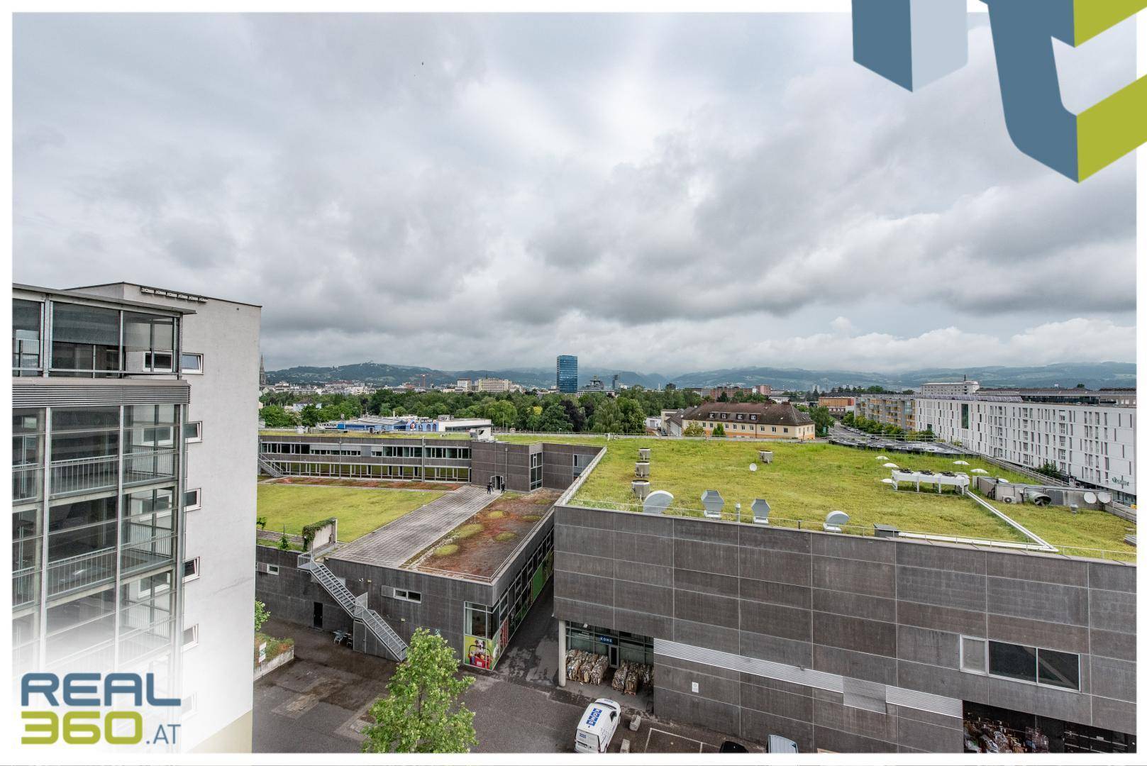 Weitblick von einem oberen Stockwerk über begrünte Dächer und die Stadtlandschaft unter bewölktem Himmel.