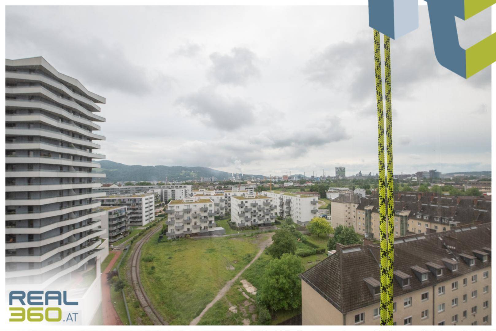 Weitläufiger Ausblick über Wohngebäude, Grünflächen und Industrieanlagen unter bewölktem Himmel.