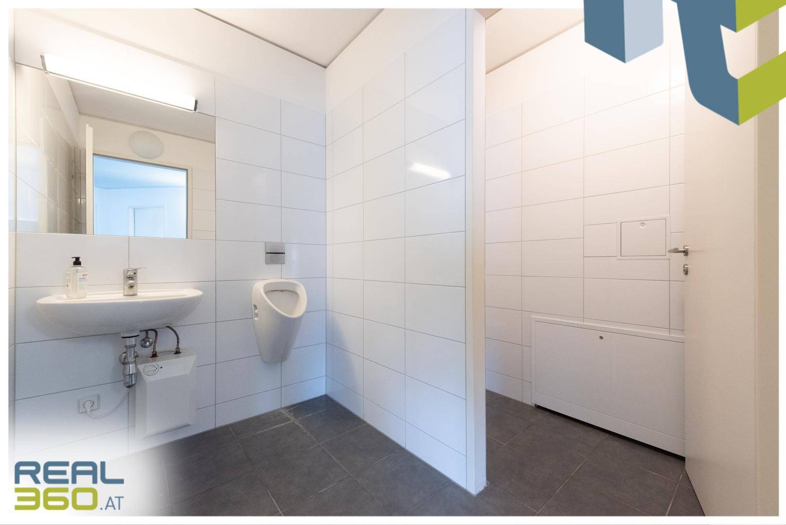 Helles Badezimmer mit weißen Fliesen, grauen Bodenfliesen, Waschbecken und einem Urinal.