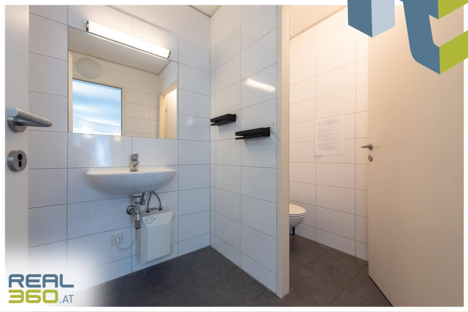 Modernes Badezimmer mit weißen Wandfliesen, grauen Bodenfliesen, Waschbecken und separater Toilette.