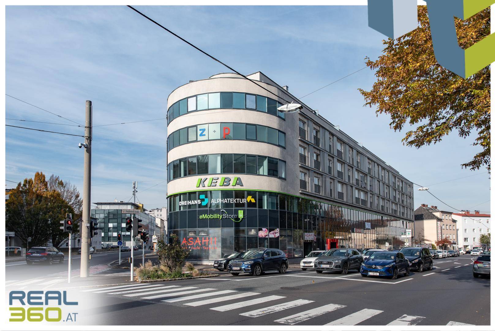 Modernes Bürogebäude an einer belebten Straßenecke mit vielen Parkplätzen und umliegenden Gebäuden.