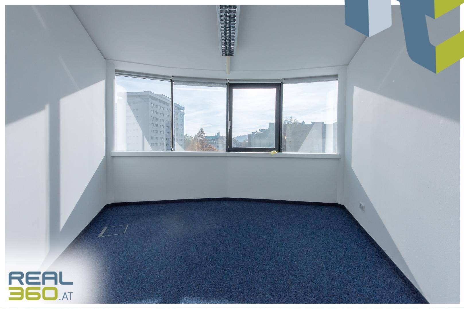 Heller Büroraum mit blauem Teppichboden und einer großen, gebogenen Fensterfront mit Stadtblick.