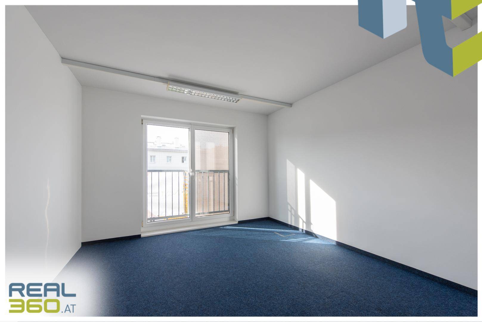 Leerer Büroraum mit blauem Teppichboden, weißen Wänden und Zugang zu einem kleinen Balkon.