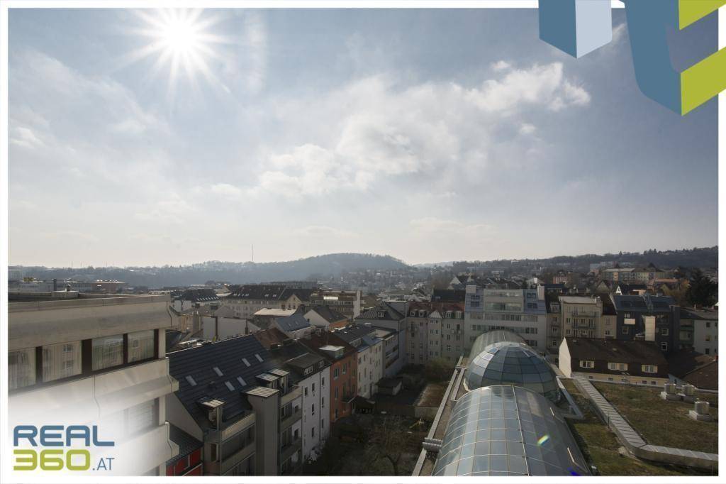 Weitläufiger Blick über die Stadt mit hügeliger Landschaft im Hintergrund unter strahlendem Sonnenschein.