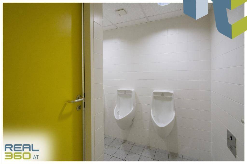 Sanitärbereich mit zwei Urinalen, weißen Fliesenwänden und einem auffälligen gelben Türblatt.