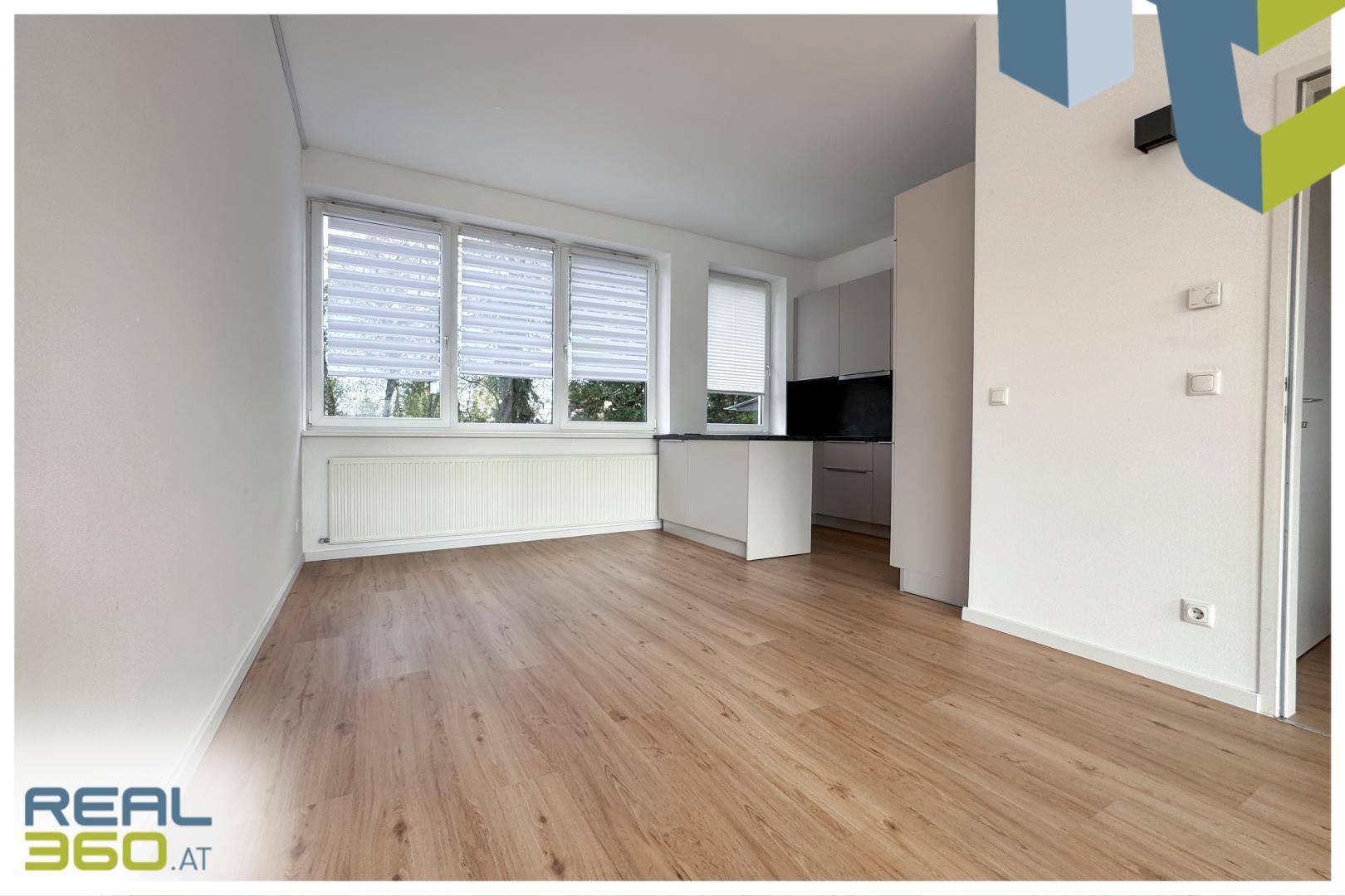 Heller Wohnbereich mit Laminatboden und Blick auf die moderne Küche im Hintergrund.