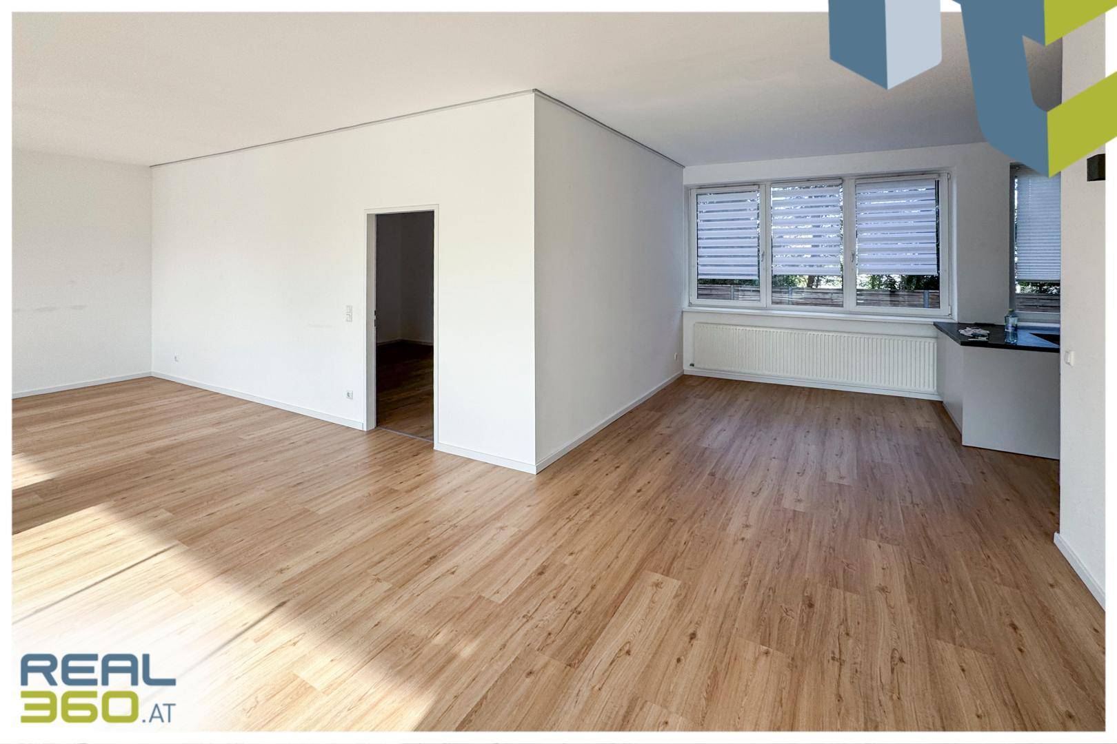 Heller Wohnbereich mit Laminatboden und Blick auf die moderne Küche im Hintergrund.