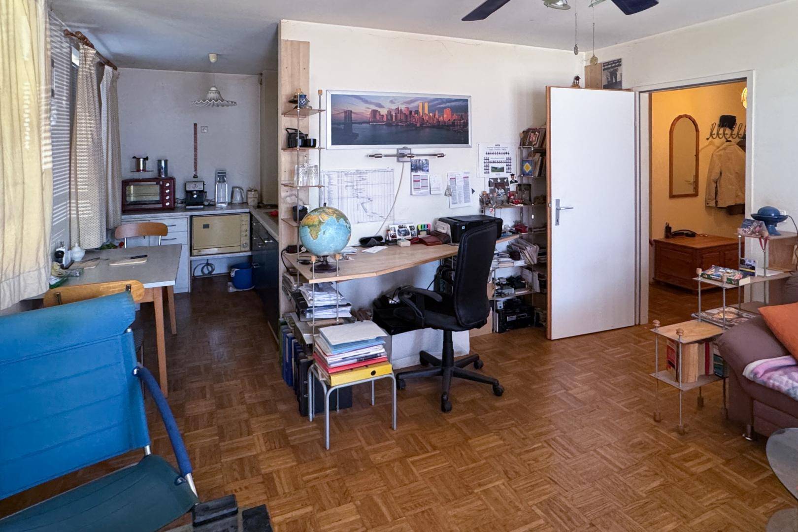 Multifunktionaler Wohnbereich mit Schreibtisch, kleiner Küche und Parkettboden.