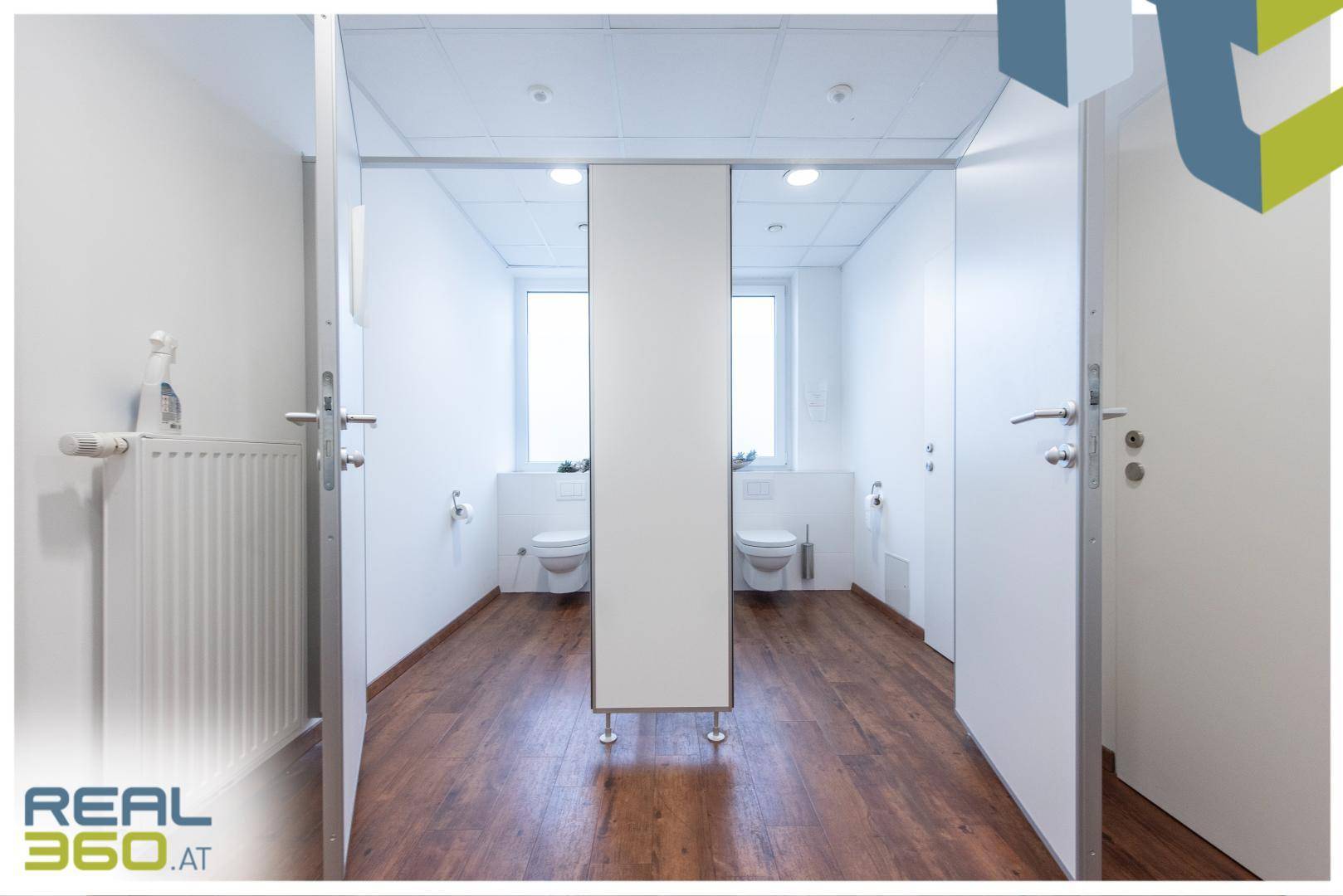 Heller Damenwaschraum mit mehreren Toilettenkabinen, Fenstern und Holzboden, sehr gepflegt.