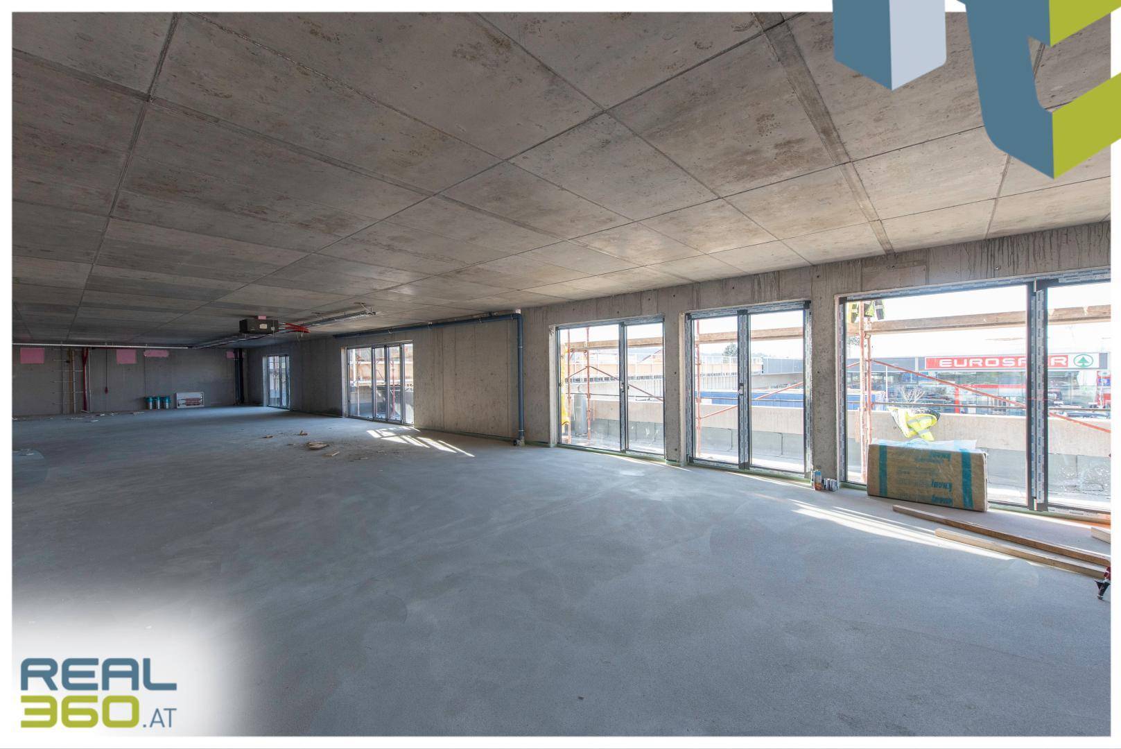 Geräumiger Rohbau-Gewerberaum mit unverputzten Wänden, Betonboden und großen Fenstern, die viel Licht hereinlassen.