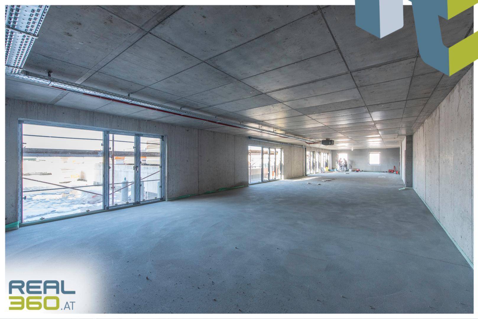Großer, offener Gewerberaum im Rohbau mit Betonboden und großen Fensterfronten für viel Tageslicht.
