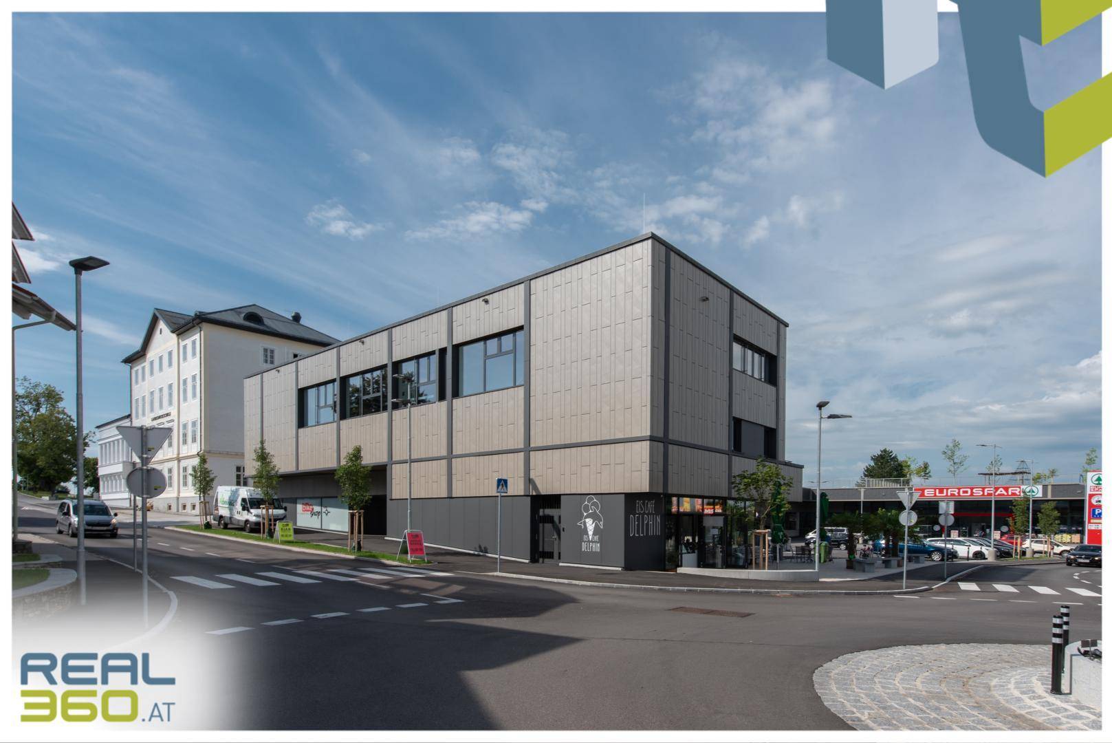 Modernes Geschäftsgebäude an einer belebten Kreuzung mit angrenzendem Supermarkt und weiteren Geschäften.