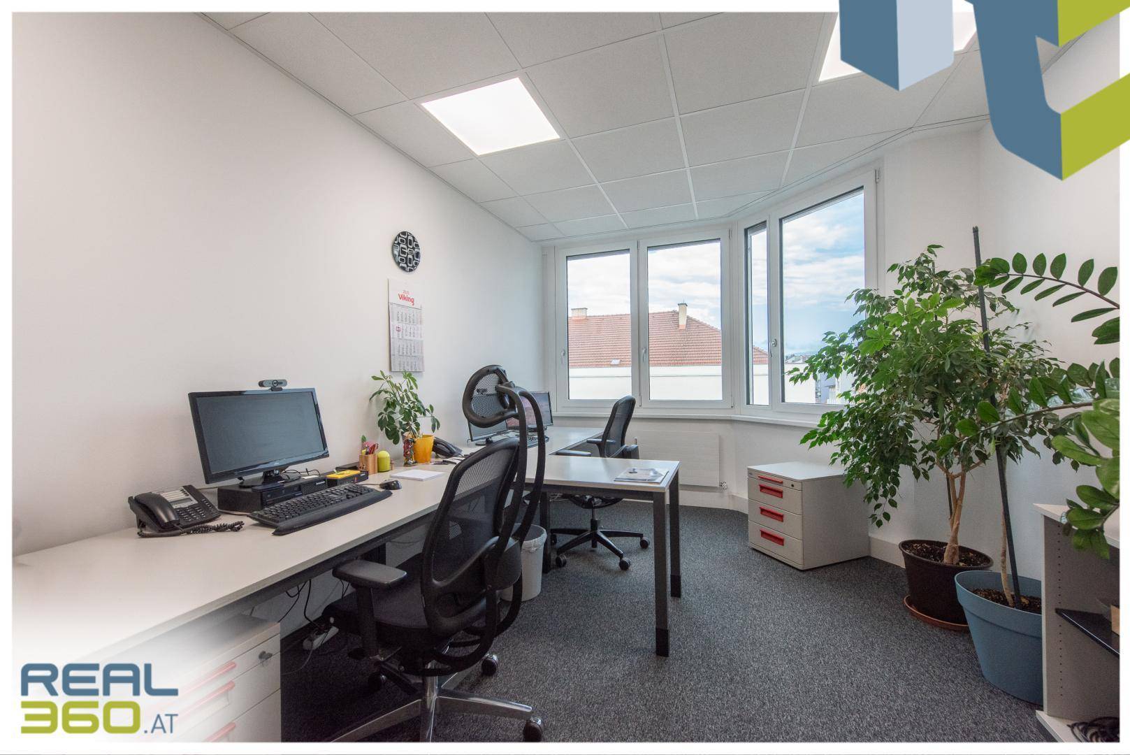 Helles Eckbüro mit mehreren Arbeitsplätzen, großen Fenstern und Blick auf die Dächer der Stadt.