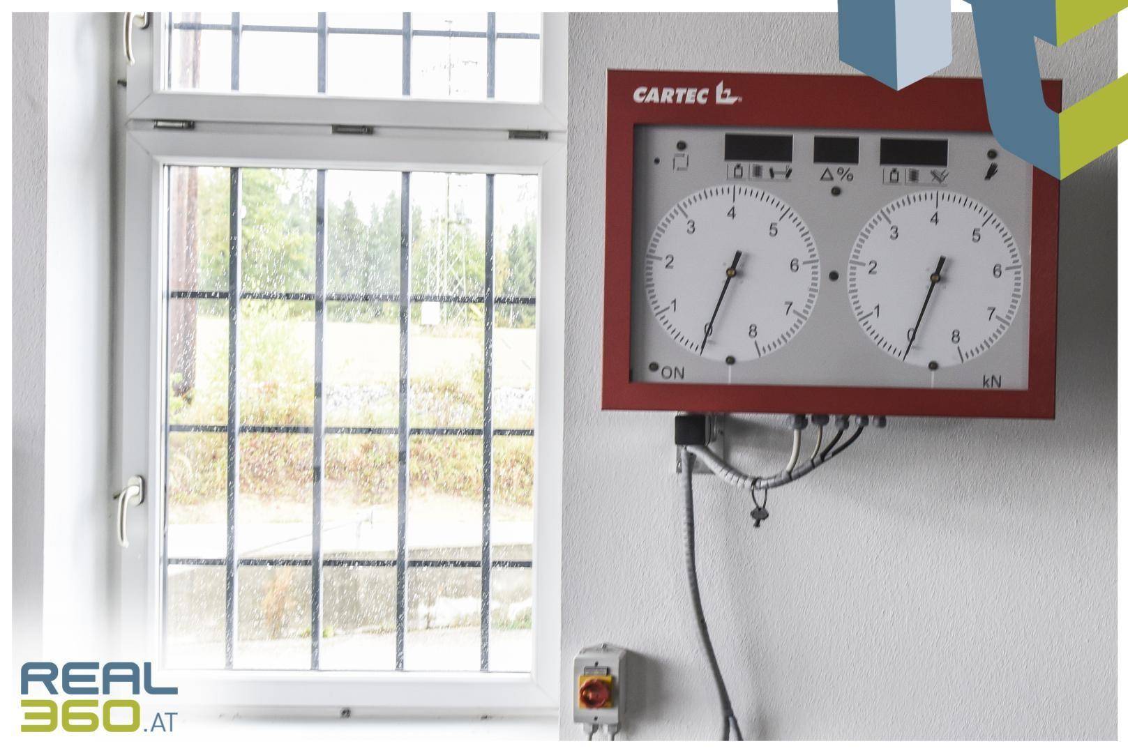 Fenster mit Gitterstäben und ein Wandgerät zur Messung oder Steuerung in einem Werkstattbereich.