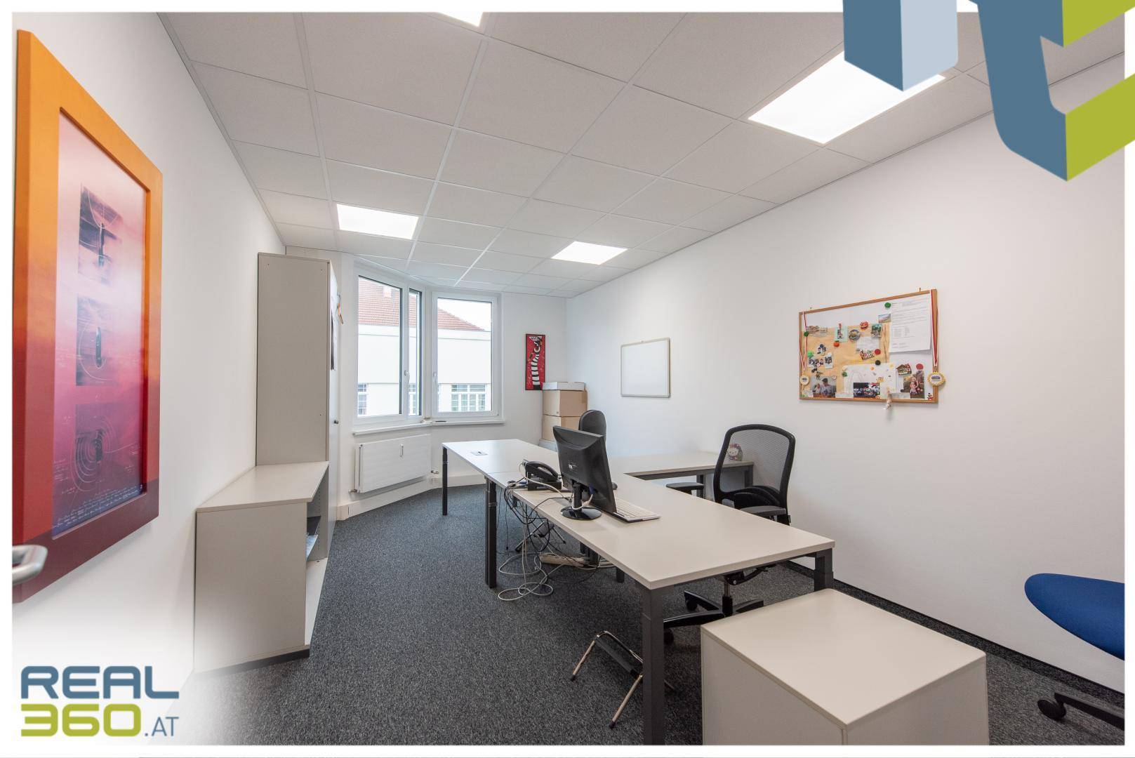 Einzelbüro mit Schreibtisch, Bürostuhl, Pinnwand und Fenstern, die viel Tageslicht hereinlassen.