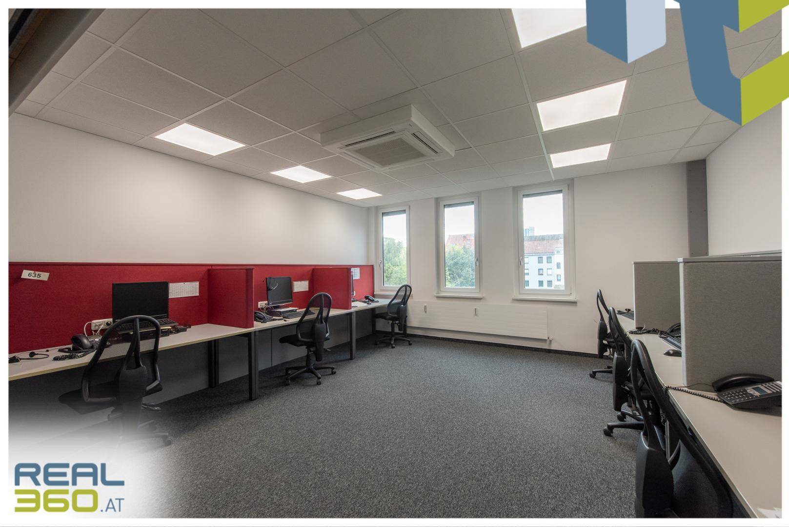 Großraumbüro mit mehreren Arbeitsplätzen, roten Trennwänden und Fenstern mit Blick nach draußen.