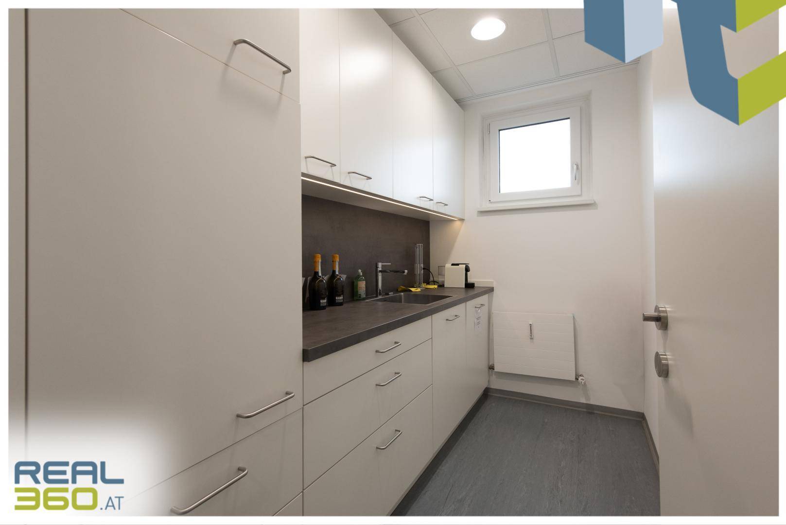 Kleine, funktionale Teeküche mit weißen Schränken, Spüle und einem Fenster für natürliche Belüftung.