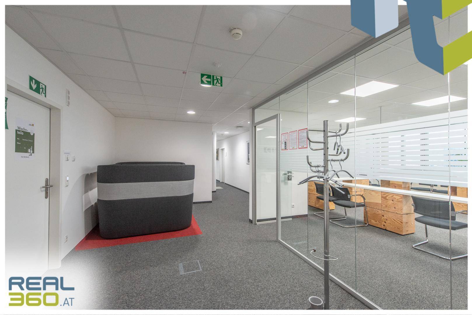 Langer Bürogang mit Teppichboden, Glaswänden zu Büros und Notausgangsschildern.
