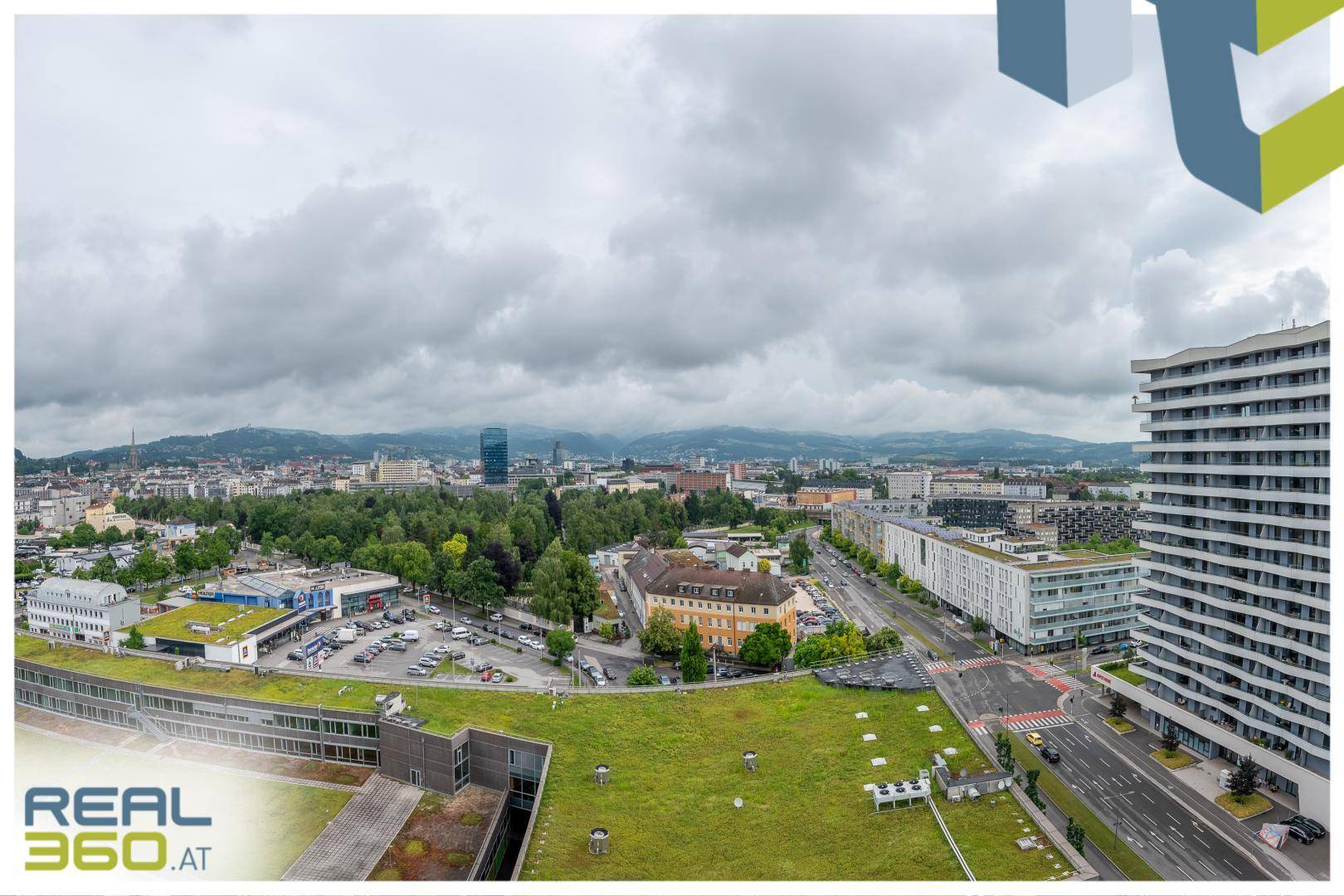 Weitläufige Luftaufnahme der Stadt mit Gebäuden, Grünflächen und Bergen im Hintergrund.