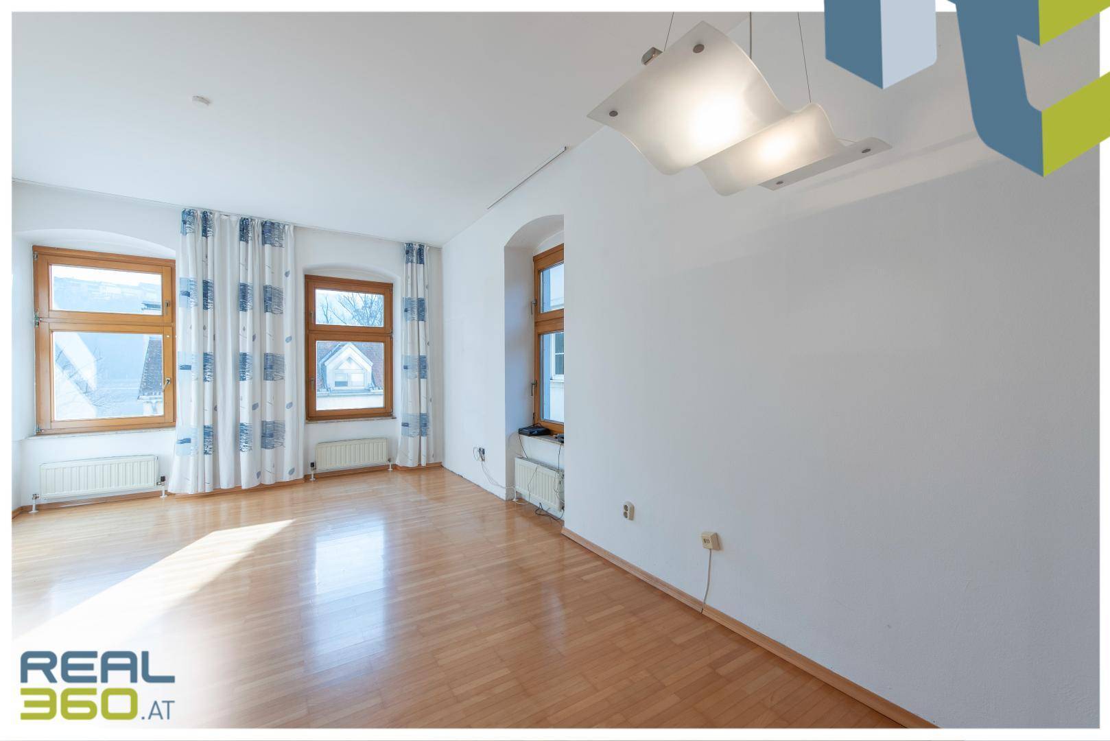 Geräumiges Wohnzimmer mit Parkettboden, großen Fenstern und moderner Deckenleuchte.