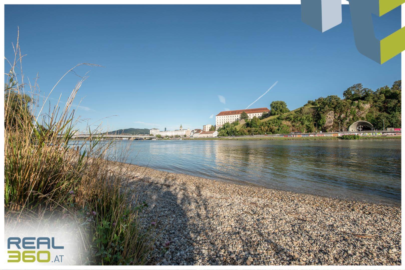 Weitläufiger Flussblick mit Kiesstrand und Gebäuden am gegenüberliegenden Ufer unter klarem Himmel.