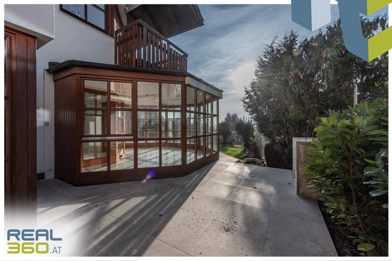 Helle Terrasse mit Fliesenboden und angrenzendem Wintergarten, umgeben von Grün.