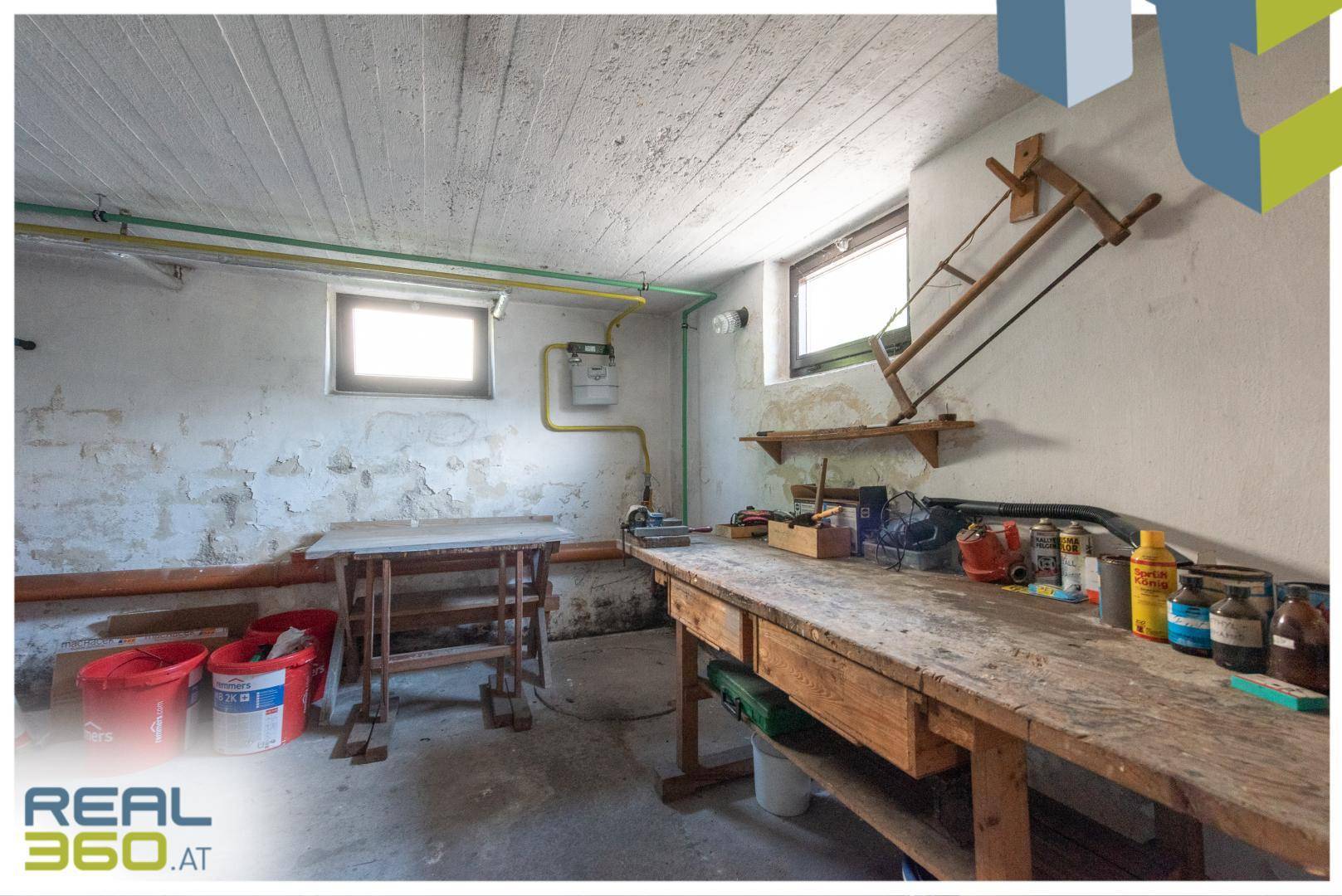 Werkstatt im Keller mit Arbeitstischen, Werkzeugen und kleinen Fenstern.
