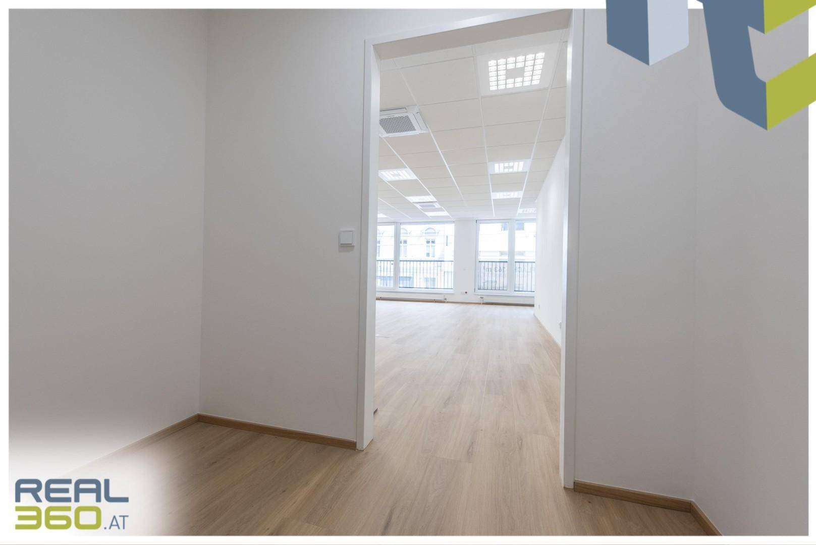 Blick von einem kleineren Raum in einen größeren, hellen Bürobereich mit Laminatboden.