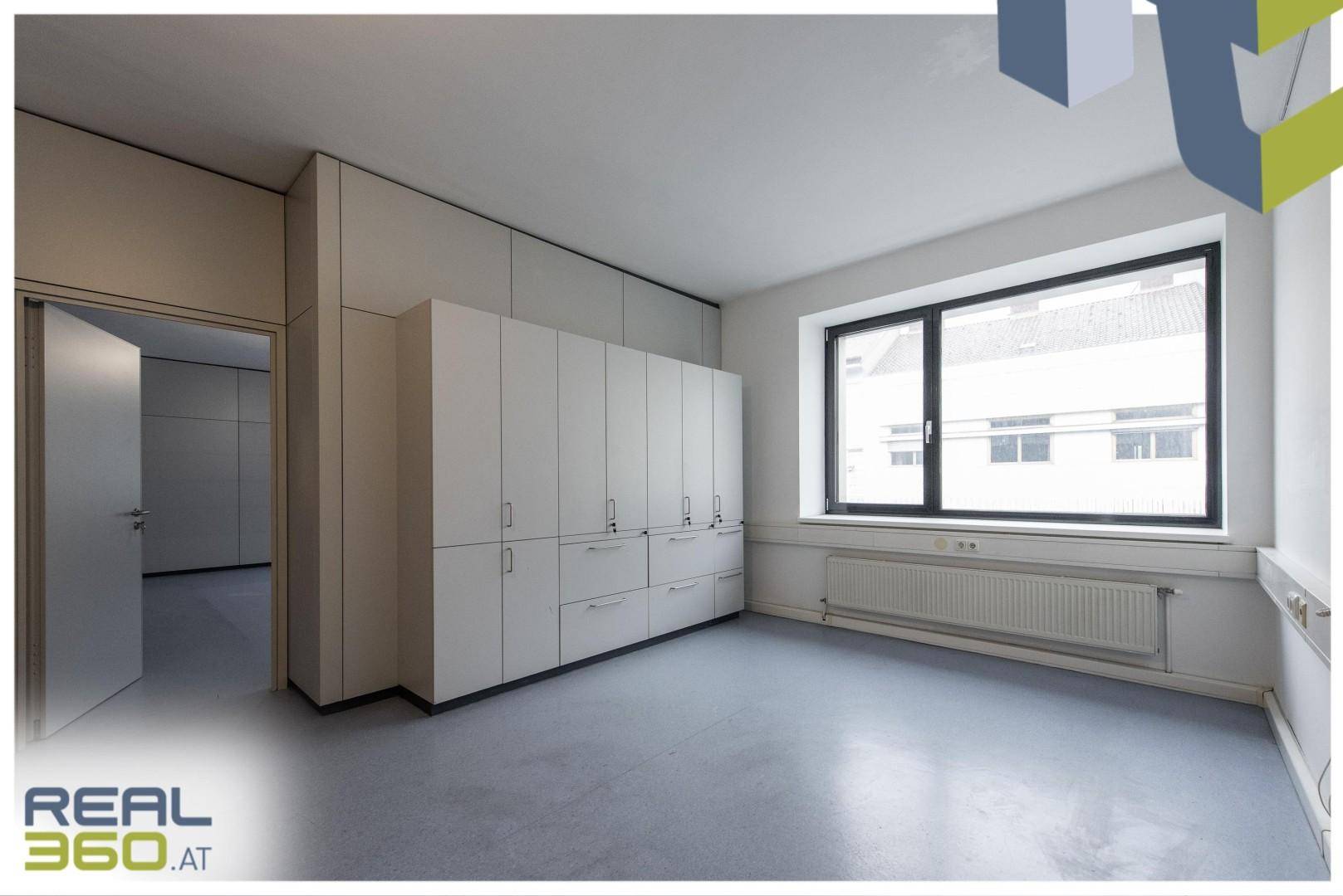 Heller Büroraum mit großem Fenster, langen weißen Schränken und grauem Bodenbelag, bereit zur individuellen Gestaltung.