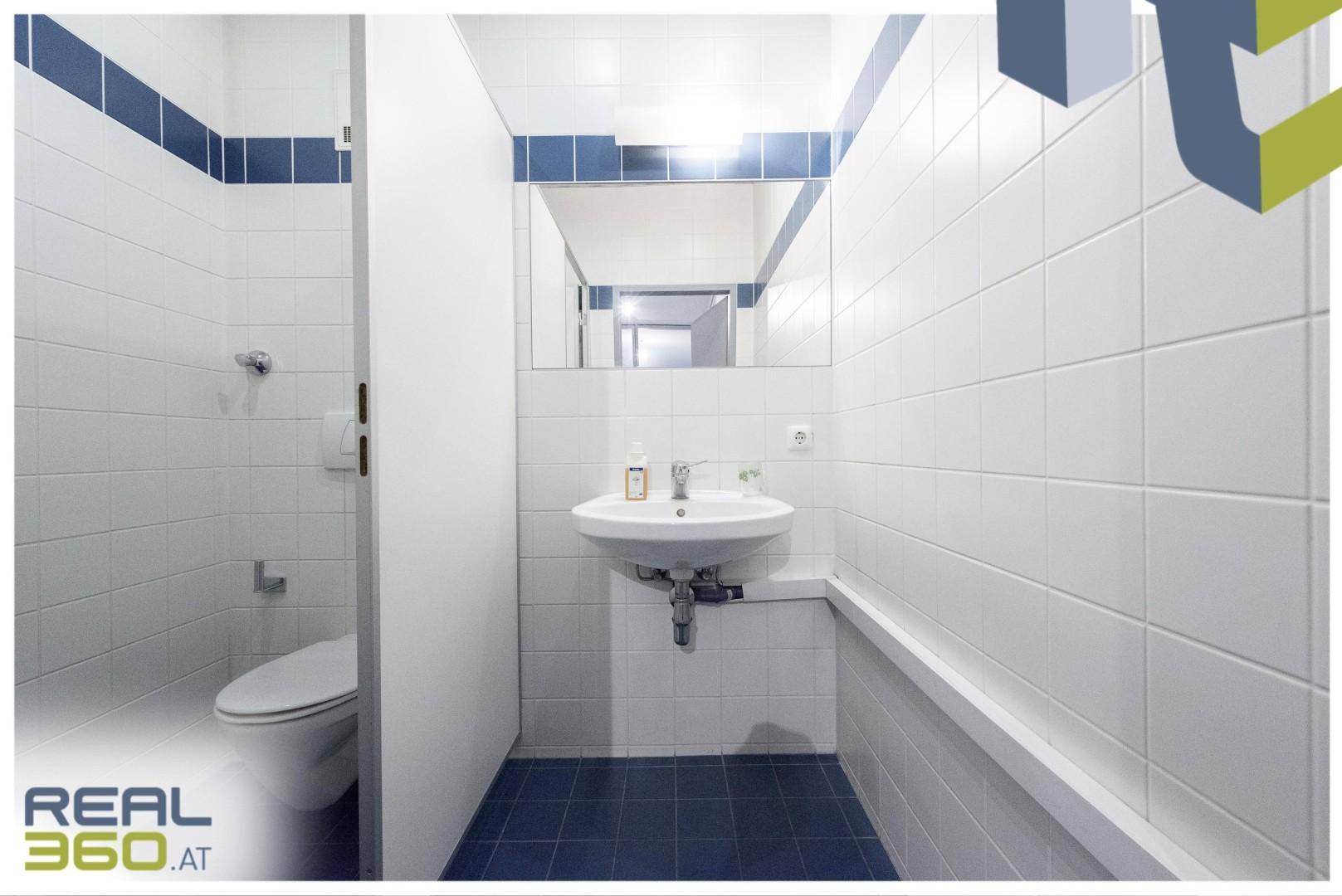 Saubere Toilette mit weißen Wandfliesen, blauem Boden, Waschbecken und Spiegel, in gutem Zustand.