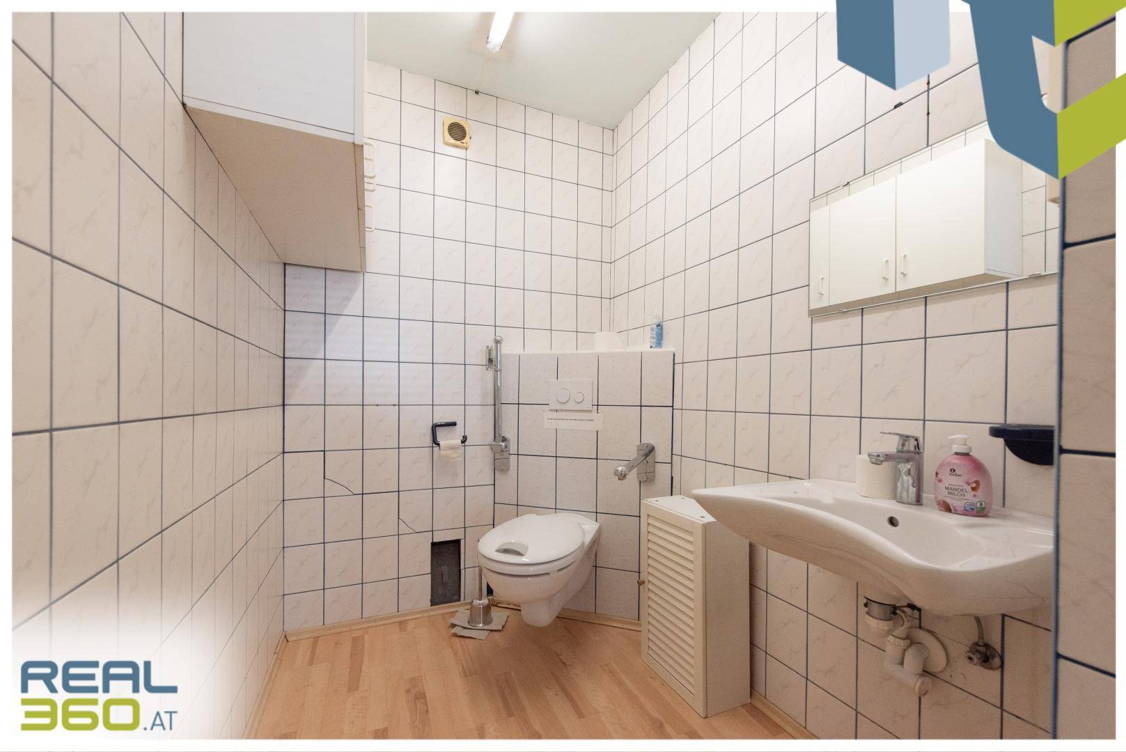 Gefliestes WC mit wandhängender Toilette, Waschbecken und Haltegriffen.