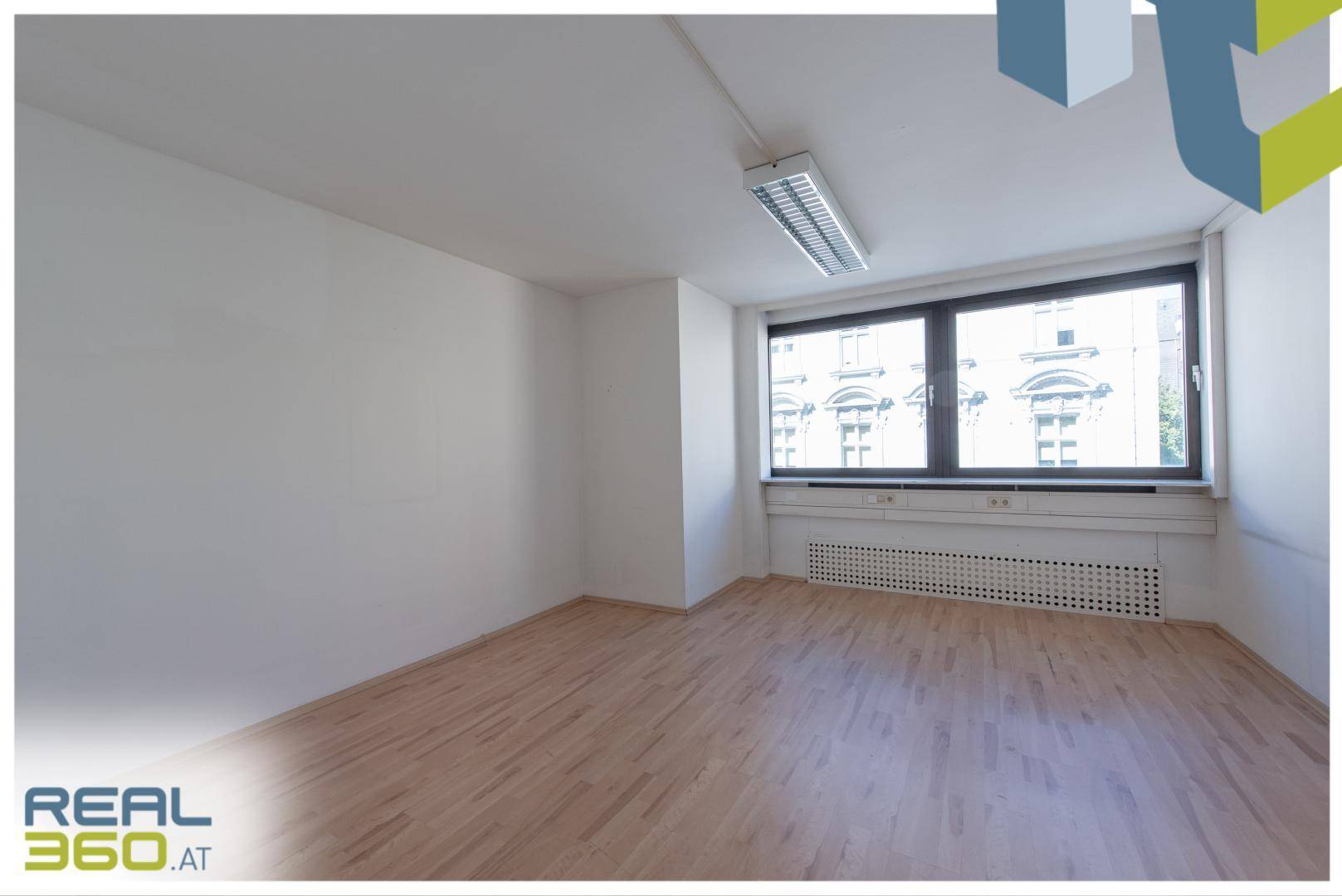 Leeres Büro mit Holzboden, großen Fenstern und Blick auf die umliegenden Gebäude.