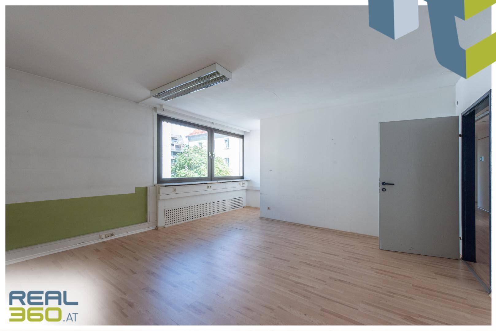 Helles Büro mit Holzboden, großen Fenstern und Blick auf Bäume und Gebäude.