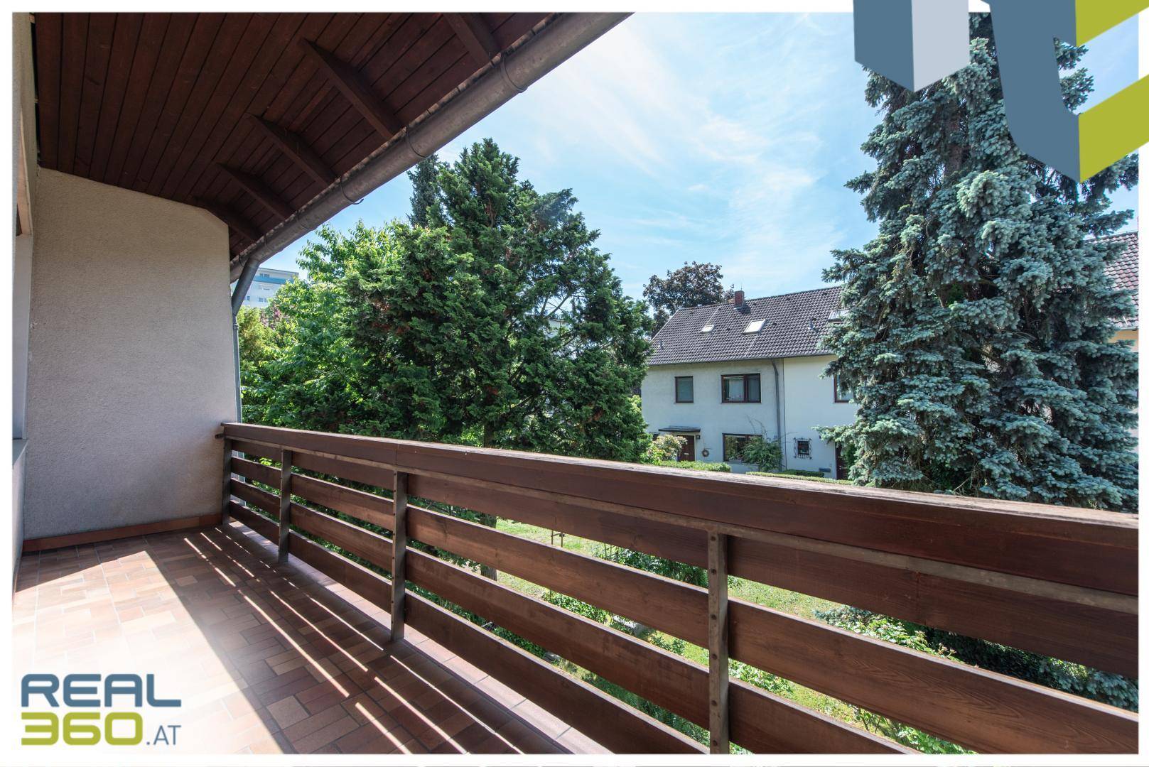 Überdachter Balkon mit Holzboden und Blick auf die umliegende grüne Landschaft und Nachbarschaft.