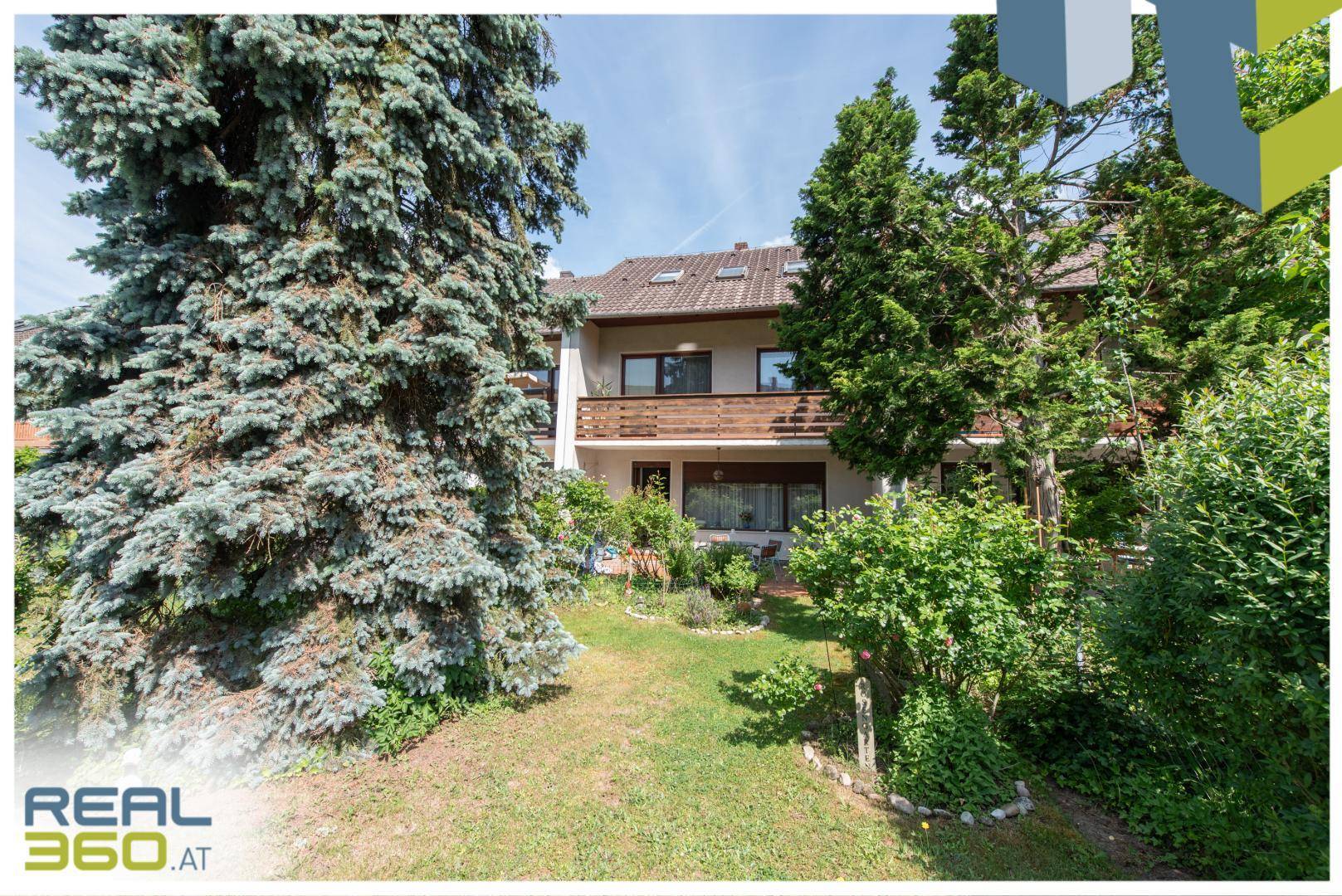 Ansicht des Hauses von der Gartenseite mit Rasenfläche und hohen Bäumen unter blauem Himmel.