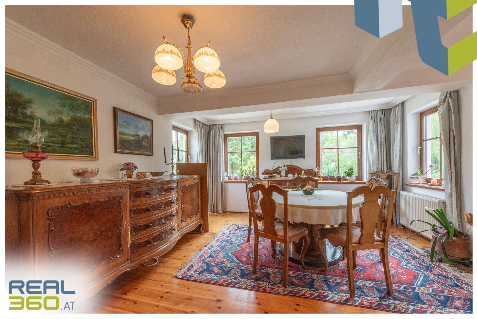 Elegantes Esszimmer mit Holzboden, antiken Möbeln und mehreren Fenstern mit Blick ins Grüne.