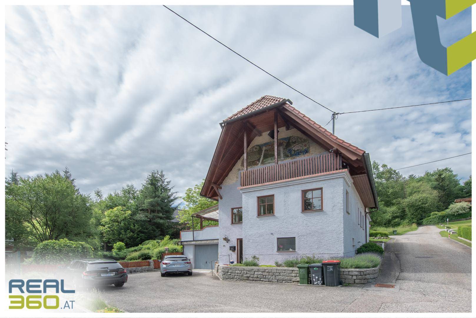 Seitenansicht des Hauses mit weißer Fassade, Holzbalkon und Zufahrt mit Parkplätzen.