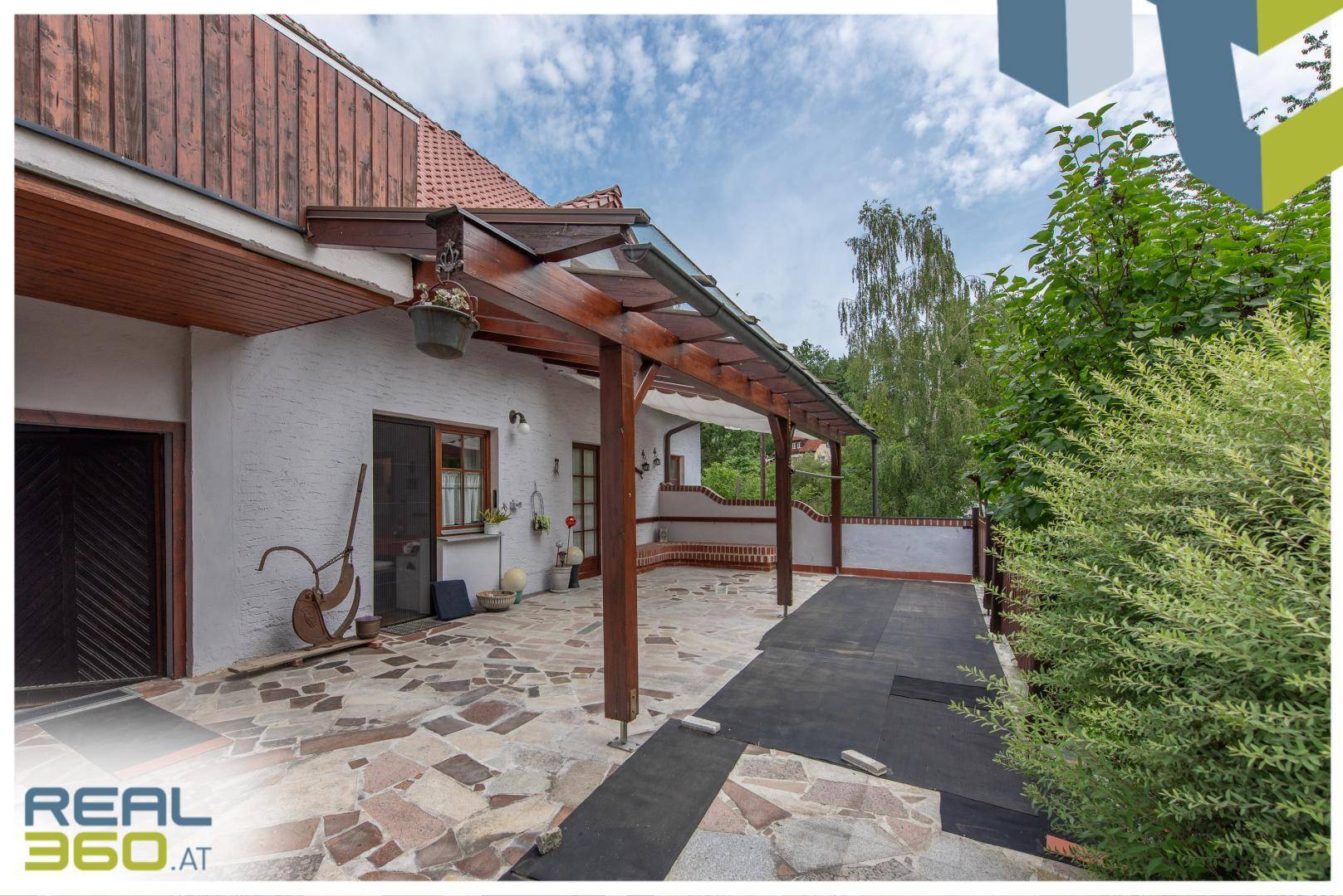 Großzügige überdachte Terrasse mit Steinplattenboden und Holzbalkenstruktur, ideal zum Entspannen.