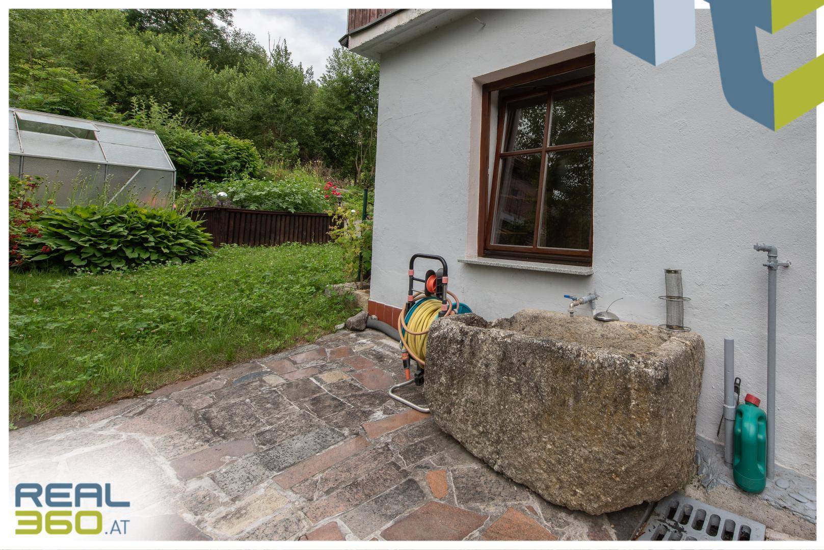 Gartenbereich mit Natursteinbrunnen, Gewächshaus und grüner Bepflanzung.