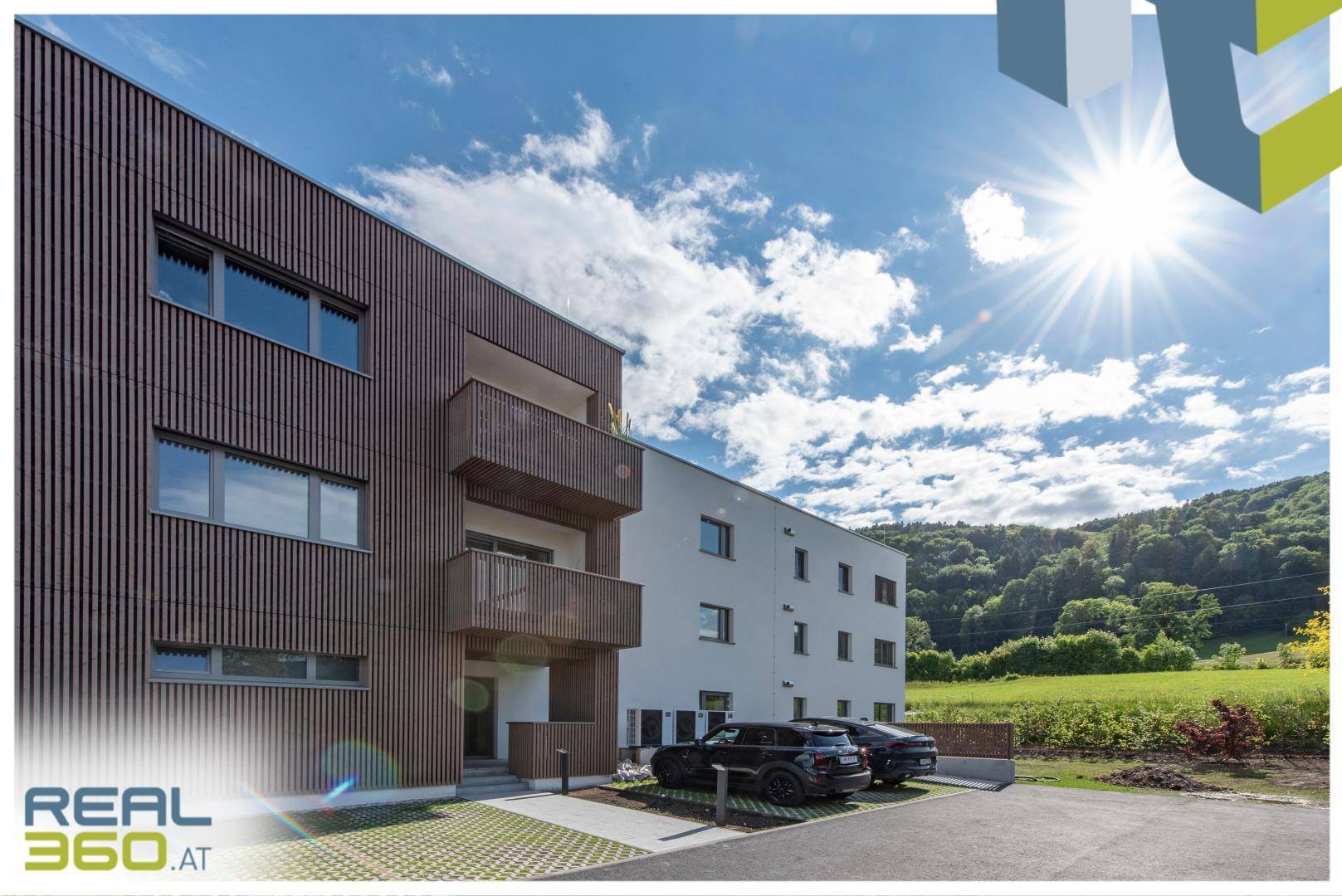 Moderne Wohnanlage mit Holzfassade und Parkplätzen vor einer grünen Hügellandschaft.
