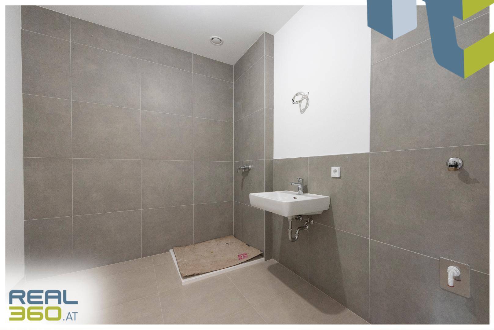 Modernes Badezimmer im Rohbau mit grauen Fliesen und Waschbeckeninstallation.