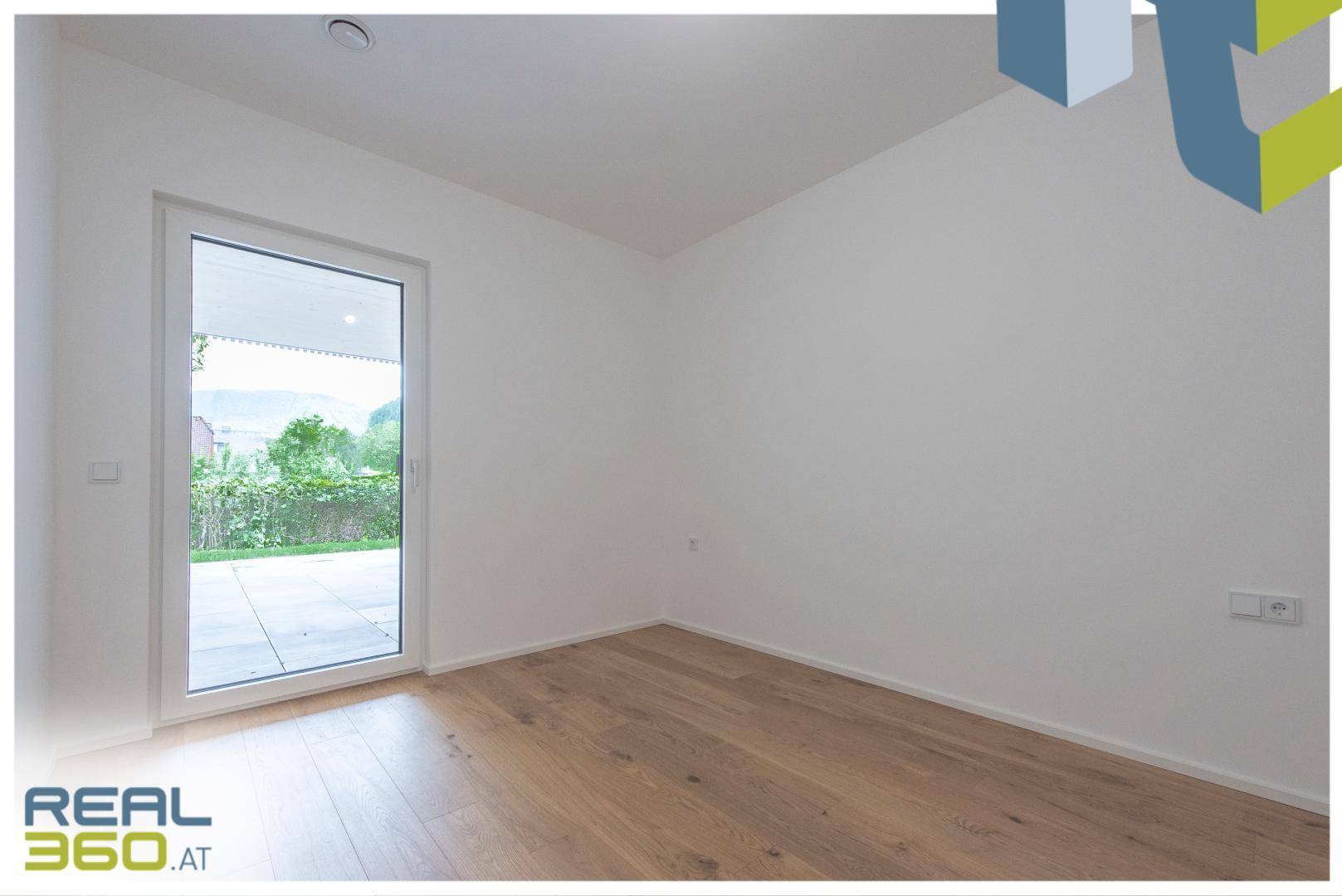 Helles Zimmer mit Holzboden und Zugang zu einem Außenbereich durch eine Glastür.