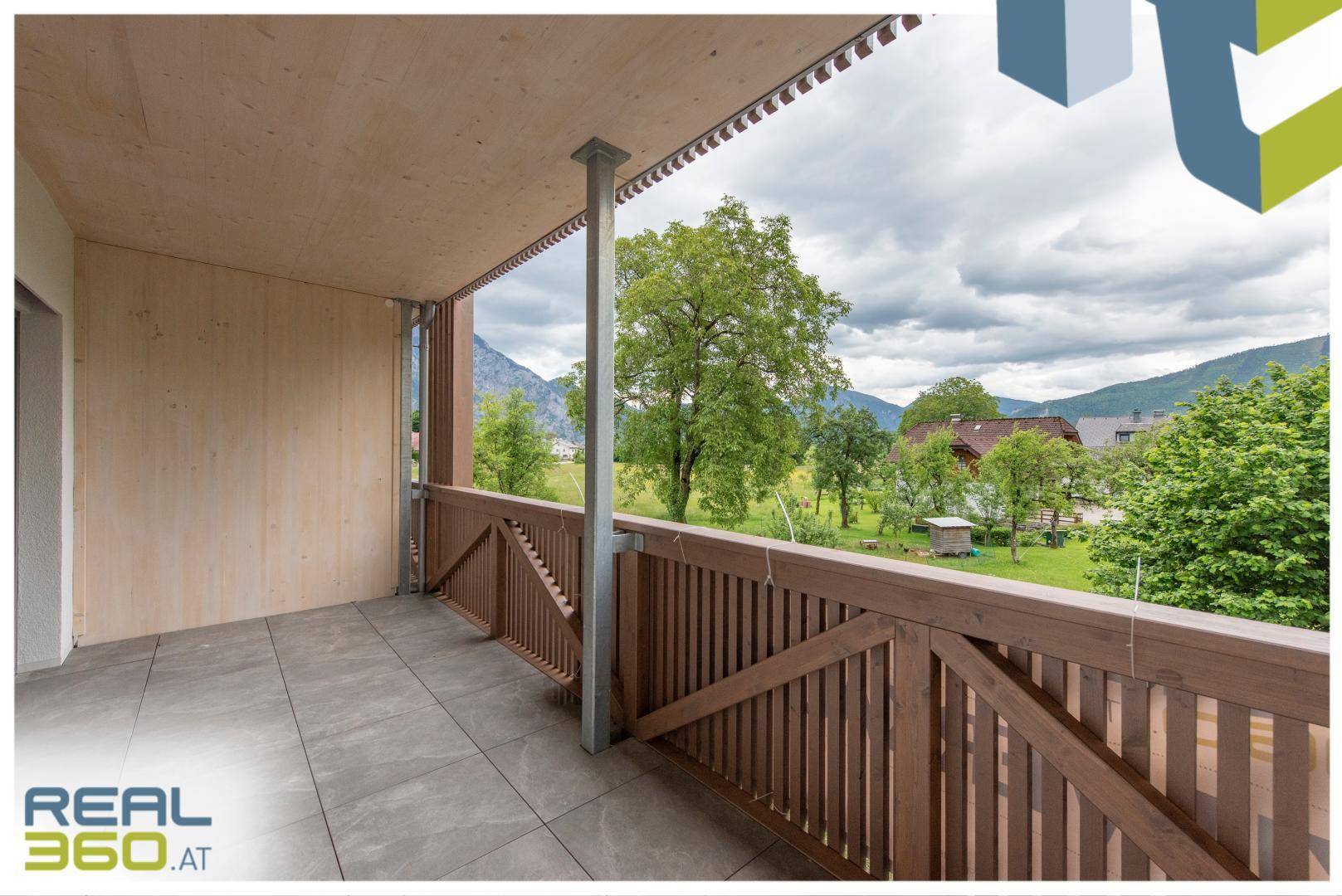 Großer Balkon mit Holzelementen und Fliesenboden, umgeben von grüner Natur und Bergblick.