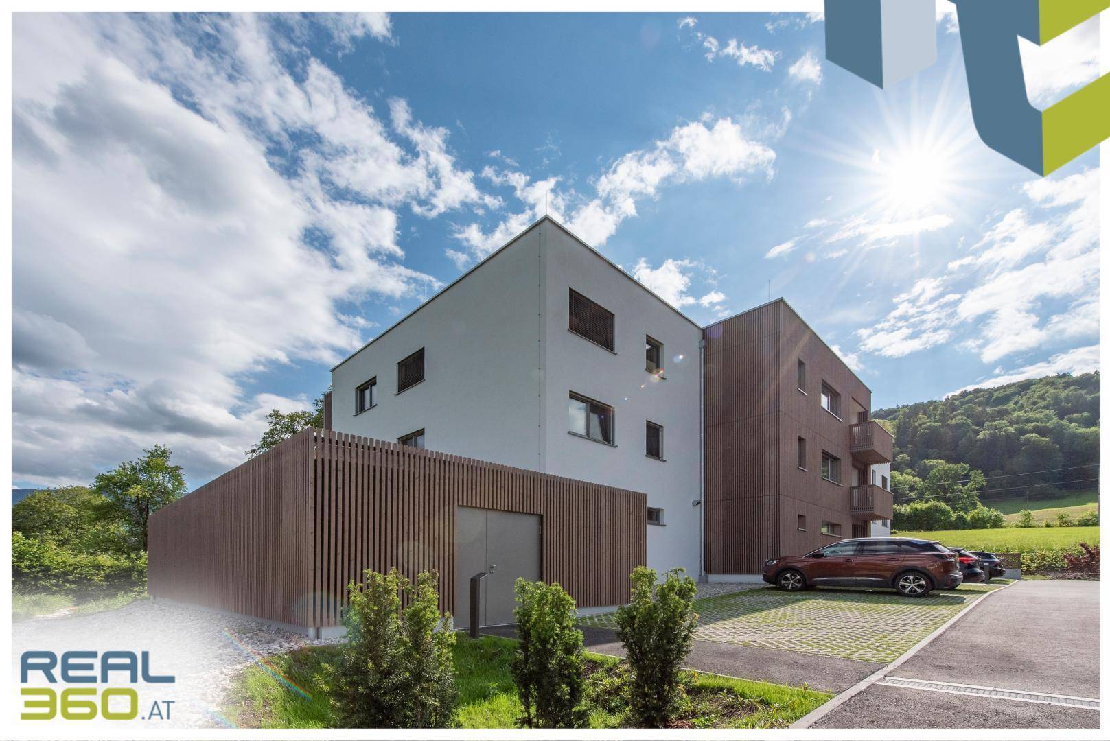 Modernes Wohngebäude mit Holzverkleidung und Parkplätzen, umgeben von Natur unter blauem Himmel.