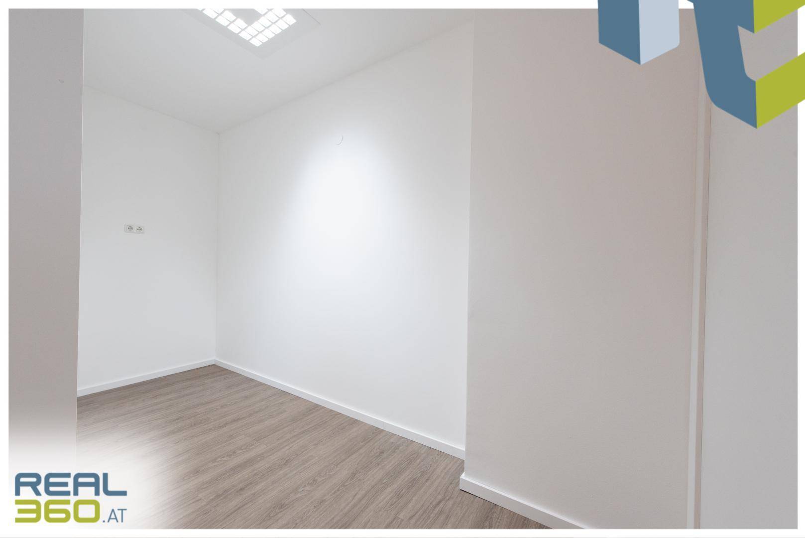 Leerer, heller Büroraum mit schlichten weißen Wänden und modernem Laminatboden.